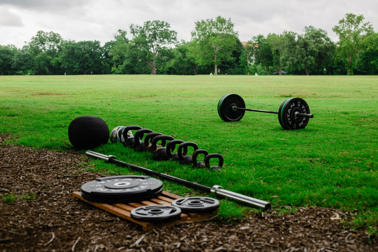 Functional fitness equipment including kettlebells, barbells, and slam balls set up on grass at YUDAE Wellness outdoor gym