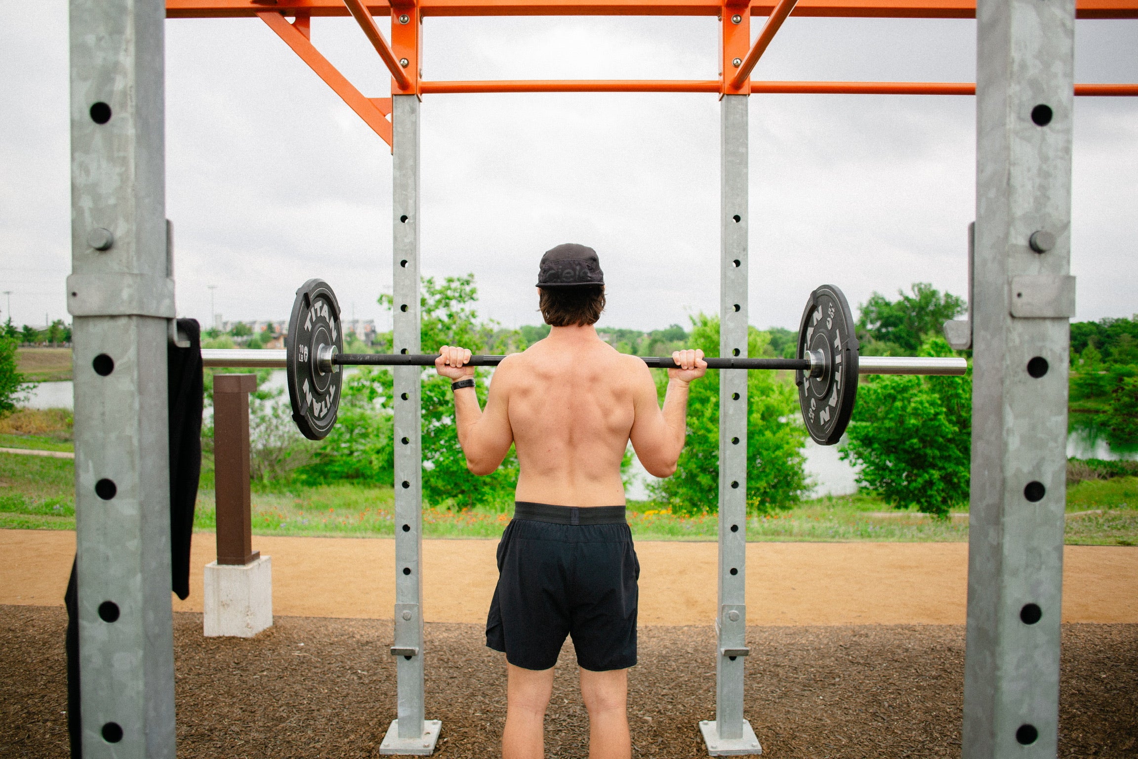 man standing at yudae wellness outdoor gym under orange calisthenics bar curling barbell wearing black shorts and a black hat